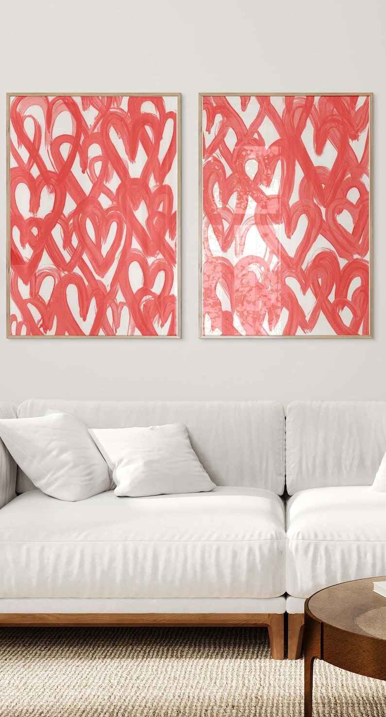May include: Two framed abstract art prints with a white background and a pattern of red hearts. The prints are hanging on a white wall above a white sofa.