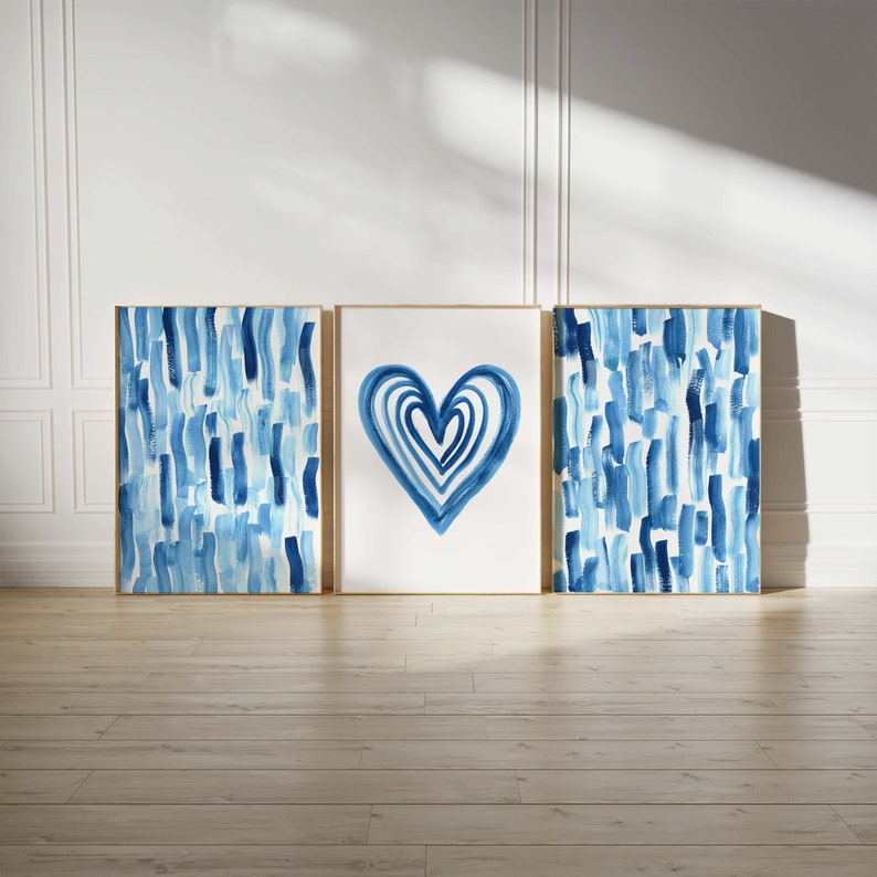 Puede incluir: Tres impresiones art&iacute;sticas enmarcadas. La impresi&oacute;n central presenta un dise&ntilde;o de coraz&oacute;n azul. Las otras dos impresiones tienen un patr&oacute;n de pinceladas azules. Las tres impresiones tienen marcos dorados y se exhiben contra una pared blanca y un suelo de madera.