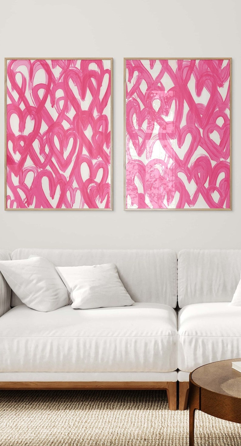 May include: Two framed abstract paintings with pink hearts on a white background. The paintings are hanging on a white wall above a white couch.