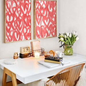 May include: A white desk with a laptop, a stack of books, a white candle, a wooden calendar showing the date 19th, a vase of white tulips, and two framed prints of pink hearts on a white background.