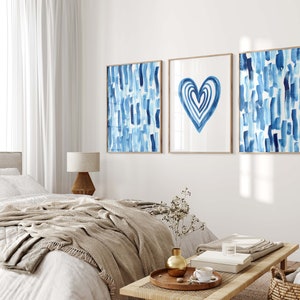 Puede incluir: Tres impresiones art&iacute;sticas enmarcadas con dise&ntilde;os de acuarela. Las impresiones presentan pinceladas azules y una forma de coraz&oacute;n azul sobre un fondo blanco. Los marcos son de color marr&oacute;n claro. La obra de arte se exhibe sobre una cama en un dormitorio.
