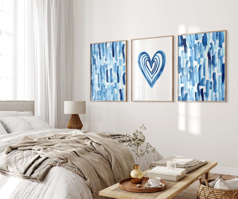 Puede incluir: Tres impresiones art&iacute;sticas enmarcadas con dise&ntilde;os de acuarela. Las impresiones presentan pinceladas azules y una forma de coraz&oacute;n azul sobre un fondo blanco. Los marcos son de color marr&oacute;n claro. La obra de arte se exhibe sobre una cama en un dormitorio.
