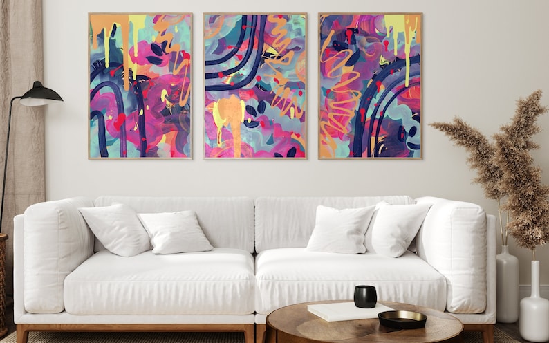 May include: Three abstract paintings in a modern living room. The paintings are framed in wood and feature a colorful, abstract design with splashes of pink, blue, yellow, and orange.