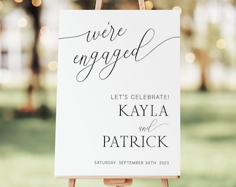 May include: A white sign on a wooden easel with the words "we're engaged" in elegant script. Below, it reads "Let's Celebrate!" followed by "Kayla and Patrick" and the date "Saturday, September 30th, 2023". The background is a blurred outdoor scene.
