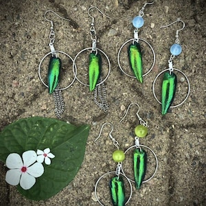 Puede incluir: Pendientes hechos a mano con alas de escarabajo verdes iridiscentes. Los pendientes tienen aros plateados, algunos con detalles de cadena o cuentas. Una hoja y flores blancas en primer plano.