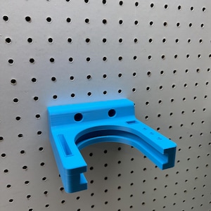 May include: A bright blue, 3D-printed pegboard tool holder. The holder has a curved design with two circular holes and two rectangular slots. It is mounted on a gray pegboard with evenly spaced holes.