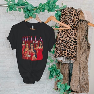 May include: Black t-shirt featuring a graphic of a woman in a red dress with the text "BELLA Goth". The shirt is hanging on a wooden hanger. Other clothing items include a leopard print top and a faux fur vest.