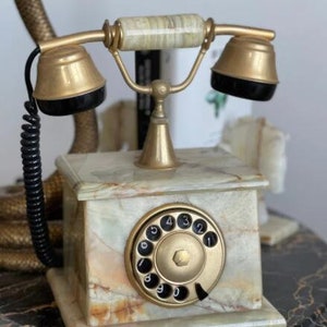 Puede incluir: Un teléfono de disco vintage con acabado dorado y base de mármol. El teléfono tiene un disco negro con números blancos y un auricular negro.