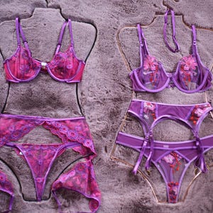 May include: Two sets of purple lingerie with floral embroidery. The first set includes a bra, suspender belt, and panty. The second set includes a bra, suspender belt, and panty. Both sets are on a light brown background.