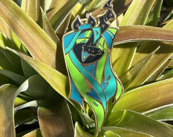 May include: A metal enamel pin featuring a stylized character with a black face, a crown, and flowing hair or fabric in shades of bright lime green and teal blue. The design is outlined in gold metal with heart-shaped cutouts and accents.