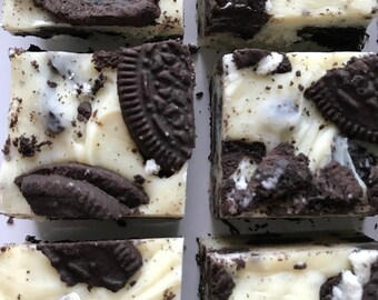 May include: A close-up of a plate of white chocolate fudge topped with crushed Oreo cookies. The fudge is cut into squares and is ready to be eaten.