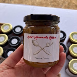 May include: A jar of pineapple chutney with a black lid, held in a hand. The label reads "Anna's Homemade Kitchen" with a spoon and fork graphic, and "Pineapple Chutney" below. Other jars are visible in the background.