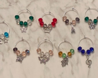May include: A collection of ten wine glass charms. Each charm features a silver ring with colorful beads and a decorative charm. Charms include a moon and star, a star with text, a crown, an Eiffel Tower, a four-leaf clover, a butterfly, a mask, a hand, and a treble clef.