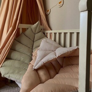 Könnte beinhalten: Ein hellgrünes, blattförmiges Kissen und ein pfirsichfarbenes, gestepptes Kissen in einem weißen Kinderbett mit einem pfirsichfarbenen Baldachin.