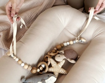 May include: A beige baby nest with a wooden and crocheted toy hanging from it. The toy features a whale, turtle, starfish, and wooden rings.