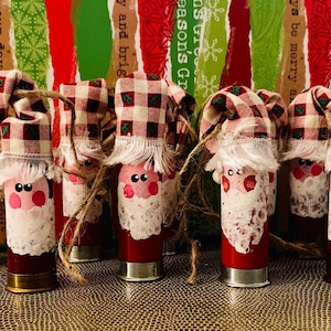 May include: A collection of red shotgun shell Christmas ornaments, each hand-painted to resemble Santa Claus. They feature white beards, pink cheeks, and plaid fabric hats, tied with twine. The background has a festive Christmas theme.