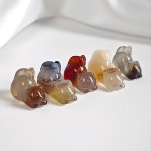 Op de afbeelding: Een verzameling van zeven kleine, gebeeldhouwde stenen berenbeeldjes in verschillende kleuren, waaronder bruin, rood en crème. De beren zijn in een rij gerangschikt, elk met een uniek patroon en tint. De achtergrond is een witte, gedrapeerde stof.