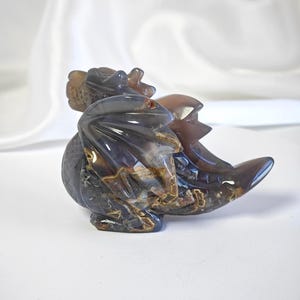 Op de afbeelding: Een uit steen gesneden drakenfiguur met een gepolijst, doorschijnend oppervlak met diepbruine, grijze en amberkleurige banden. Het wezen is afgebeeld in een zittende houding met gevouwen vleugels, een gestructureerde staart en schubdetails.