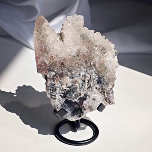May include: A large, raw crystal cluster with clear, pointed formations, displaying shades of white, gray, and hints of red and green. The crystal is mounted on a black metal stand with a circular base, set against a white background.