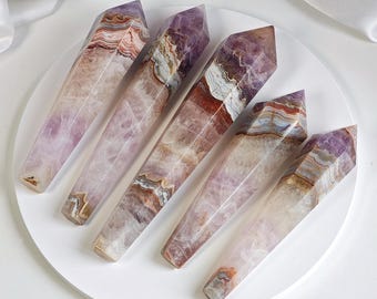 May include: Five amethyst crystal points with varying shades of purple, brown, and white. The crystals are arranged on a white circular surface. The crystals have a polished, smooth appearance and are cut into a pointed shape.