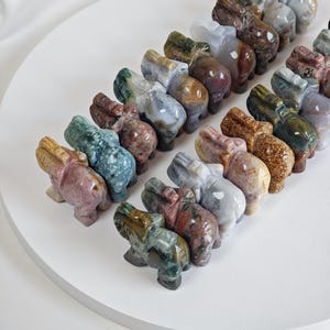 Könnte beinhalten: Eine Sammlung kleiner, geschnitzter Elefantenfiguren in verschiedenen Farben, darunter Grün, Braun, Rosa und Grau. Die Elefanten sind in ordentlichen Reihen auf einer weißen Oberfläche angeordnet und zeigen ihre detaillierte Handwerkskunst und einzigartigen Muster.