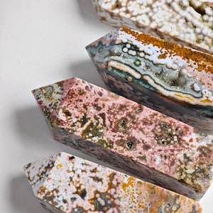 Puede incluir: Cuatro obeliscos de piedras preciosas pulidas en tonos rosa, marrón, blanco y verde. Los obeliscos tienen forma rectangular con una parte superior puntiaguda y un patrón natural moteado. Las piedras están dispuestas en diagonal sobre una superficie blanca.