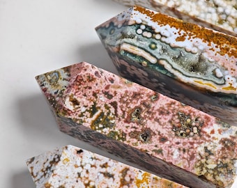 May include: Four polished gemstone obelisks in shades of pink, brown, white, and green. The obelisks have a rectangular shape with a pointed top and a speckled, natural pattern. The gemstones are arranged diagonally on a white surface.