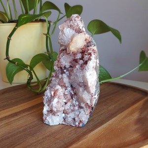 May include: A cluster of white and pink crystals with a large, light orange crystal in the center. The crystals are set in a dark gray, rock-like base. The cluster is sitting on a wooden surface.