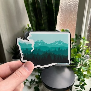 May include: A sticker of the state of Washington with a silhouette of mountains and trees in shades of green and blue. The sticker has a white outline and the text "PATHFINDER-GOODS.COM" at the bottom.