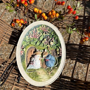 May include: A white oval ceramic plate with a raised 3D design of two mice in a garden setting. The mice are wearing clothing and are standing on a path. The background features a small door, flowers, and leaves.