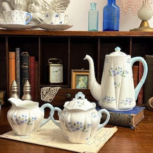 Puede incluir: Un juego de té vintage con un diseño floral blanco y azul claro. El juego incluye una tetera, azucarero y jarra de crema, todos con asas azules claras. El juego se exhibe sobre una superficie de madera con libros antiguos y objetos decorativos.