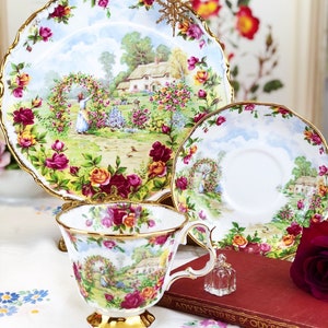 K&ouml;nnte beinhalten: Ein Set aus Vintage-Teetassen und Untertassen mit einem Blumenmuster. Die Tassen und Untertassen haben einen zarten goldenen Rand und eine charmante Cottage-Szene mit einer Frau, die sich um einen Rosengarten k&uuml;mmert. Das Set wird auf einer wei&szlig;en Tischdecke mit einem Buch und einer kleinen Flasche pr&auml;sentiert.