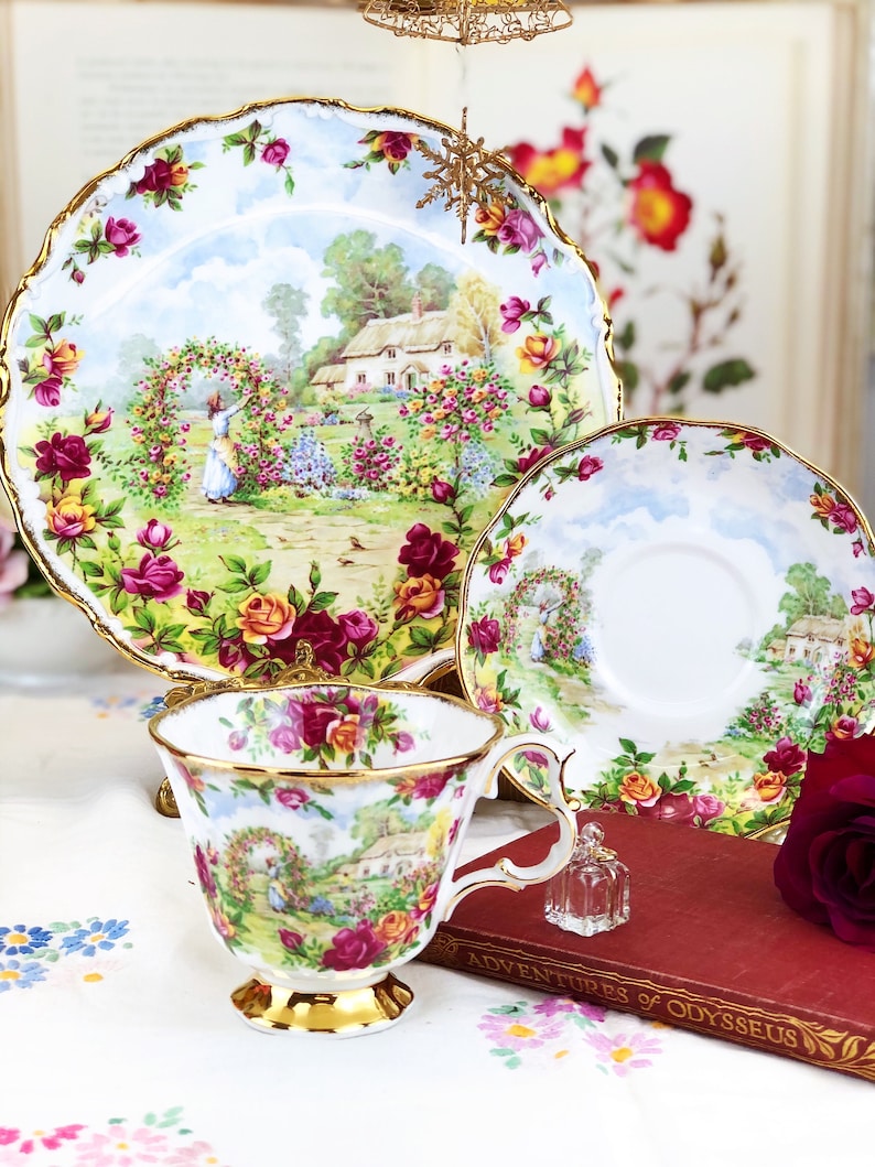 K&ouml;nnte beinhalten: Ein Set aus Vintage-Teetassen und Untertassen mit einem Blumenmuster. Die Tassen und Untertassen haben einen zarten goldenen Rand und eine charmante Cottage-Szene mit einer Frau, die sich um einen Rosengarten k&uuml;mmert. Das Set wird auf einer wei&szlig;en Tischdecke mit einem Buch und einer kleinen Flasche pr&auml;sentiert.