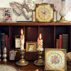 Peut inclure: Une présentation sur le thème vintage avec une statue de chérubin, des horloges anciennes et des bougies allumées. Deux bougies avec des motifs floraux sont dans des supports en laiton. Une horloge carrée avec un motif floral est visible.
