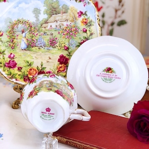 K&ouml;nnte beinhalten: Eine wei&szlig;e Teetasse und Untertasse mit einem Blumenmuster. Die Teetasse hat einen goldenen Rand und die Untertasse einen goldenen Rand und einen gewellten Rand. Die Teetasse und Untertasse sind mit einer Szene eines H&auml;uschens in einem Garten mit Rosen verziert. Die Untertasse tr&auml;gt den Text "A celebration of the Old Country Roses of the English Garden" auf der Unterseite. Die Teetasse und Untertasse stehen auf einer wei&szlig;en Tischdecke mit einem Blumenmuster.