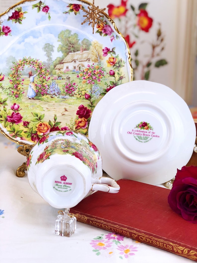 K&ouml;nnte beinhalten: Eine wei&szlig;e Teetasse und Untertasse mit einem Blumenmuster. Die Teetasse hat einen goldenen Rand und die Untertasse einen goldenen Rand und einen gewellten Rand. Die Teetasse und Untertasse sind mit einer Szene eines H&auml;uschens in einem Garten mit Rosen verziert. Die Untertasse tr&auml;gt den Text "A celebration of the Old Country Roses of the English Garden" auf der Unterseite. Die Teetasse und Untertasse stehen auf einer wei&szlig;en Tischdecke mit einem Blumenmuster.