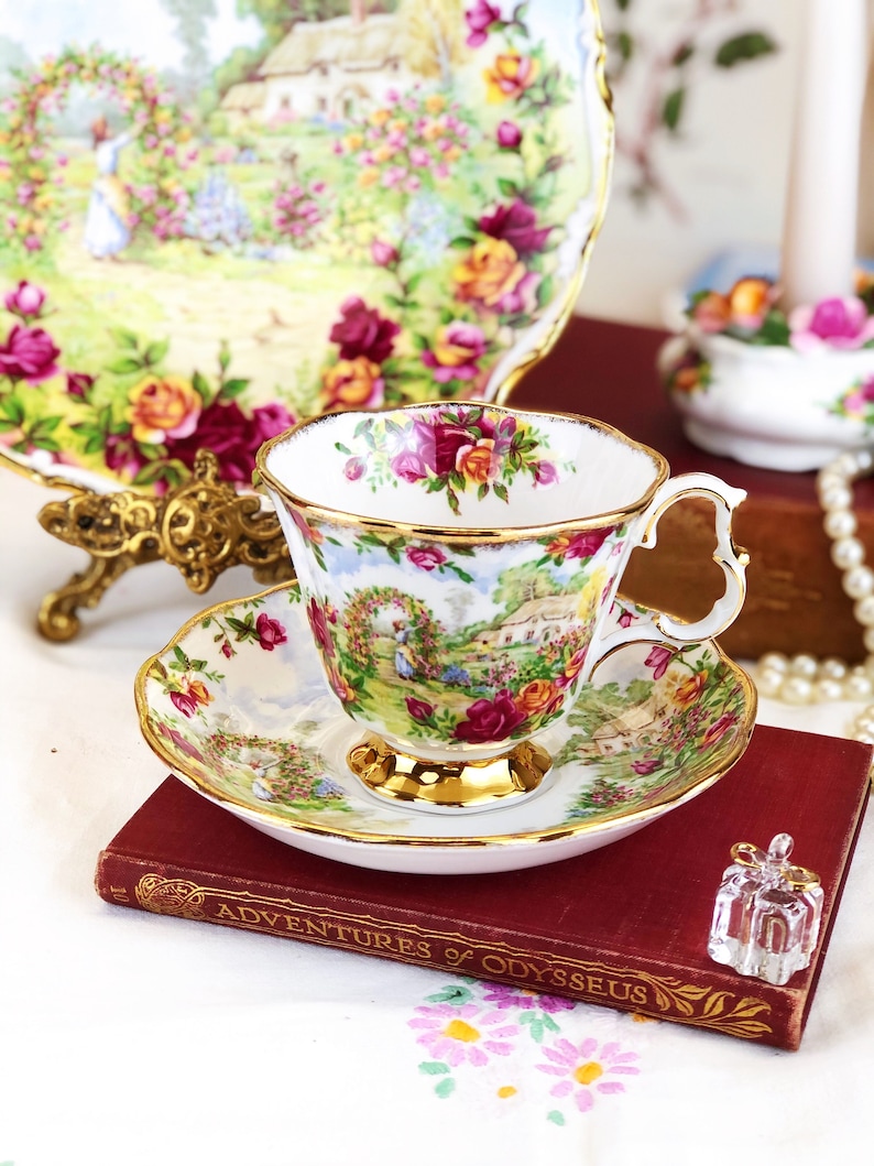 K&ouml;nnte beinhalten: Eine wei&szlig;e Teetasse und Untertasse mit floralem Muster und goldener Verzierung. Die Teetasse und Untertasse stehen auf einem roten Buch mit dem Titel "Die Abenteuer des Odysseus".
