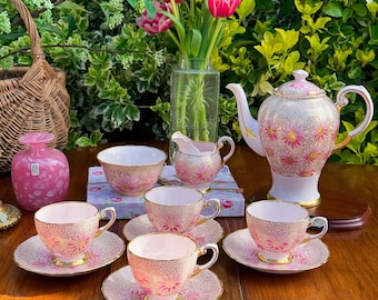 May include: A delicate tea set featuring pink floral patterns, comprising teacups, saucers, a teapot, a creamer, and a sugar bowl. The set is displayed on a wooden table, complemented by a vase of pink tulips and a wicker basket. Gold detailing adorns the edges.