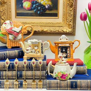 Puede incluir: Una colección de teteras vintage de diversas formas y diseños, incluyendo una tetera con forma de chimenea, una tetera con forma de librería y una tetera con forma de sillón. Las teteras están dispuestas sobre una pila de libros con detalles dorados. También hay varias cucharaditas doradas sobre los libros.