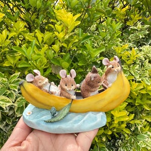 Könnte beinhalten: Eine skurrile Figur mit vier Mäusen in einem gelben Bananenboot. Das Boot ruht auf einer hellblauen Basis mit einem kleinen grünen Blatt und einem winzigen Fisch. Die Mäuse haben braunes Fell und rosa Ohren.
