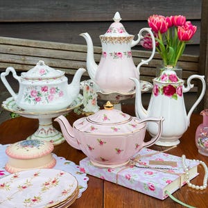 May include: A collection of vintage teacups and teapots with floral designs. The teacups and teapots are white and pink with gold trim. A stack of plates, a decorative box, and a journal are also on the table. A vase of pink tulips is in the background.