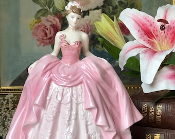 May include: A porcelain figurine of a woman in a pink gown. The dress has a layered design with intricate detailing. The figurine has a delicate face, a pearl necklace, and a floral headpiece. The background features a floral painting and a pink lily.