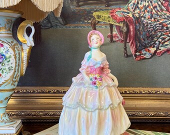 以下が含まれることがあります： ピンクと黄色のドレスを着た女性の磁器製フィギュア。花束を持っています。フィギュアはボンネットをかぶり、「LE JARDIN DES ANNIVERSAIRES」というタイトルの本の上に立っています。背景には絵画とランプがあります。