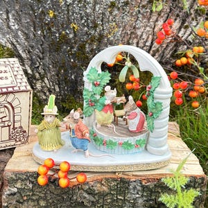 Może przedstawiać: Zestaw ceramicznych figurek przedstawiający cztery antropomorficzne myszy ubrane w stroje w stylu wiktoriańskim. Dwie myszy stoją na podstawie, a dwie pozostałe grają muzykę na podwyższeniu pod białym łukiem ozdobionym liśćmi ostrokrzewu.