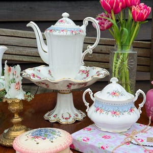May include: A vintage tea set featuring a white teapot, sugar bowl, and cake stand, all adorned with floral patterns and gold accents. A vase of pink tulips is also present. The items are displayed on a wooden table.