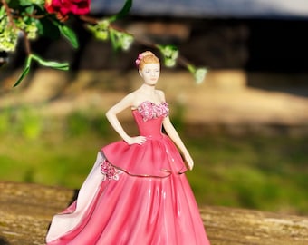 May include: A porcelain figurine of a woman in a pink strapless gown with a full skirt. The dress has floral accents and gold trim. The woman has blonde hair and is posed with one hand on her hip. A bouquet of pink and red roses is in the background.