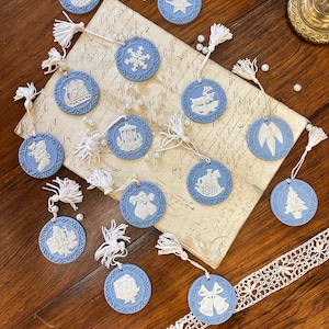 Puede incluir: Una colección de 14 adornos navideños de cerámica azul y blanca con varios diseños de temática navideña, incluyendo un muñeco de nieve, un reno, un árbol de Navidad y una campana. Los adornos están enhebrados con cinta blanca y borlas blancas.
