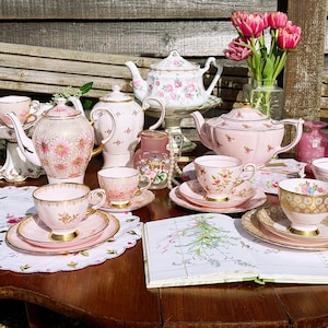 May include: A collection of pink and white porcelain teapots, teacups, and saucers featuring floral patterns and gold trim. The set includes multiple teapots, matching cups on saucers, a small frosted glass pitcher, and a book with floral illustrations.