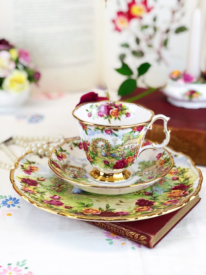 K&ouml;nnte beinhalten: Ein Vintage-Teetassen- und Untertassen-Set mit einem Blumenmuster. Tasse und Untertasse haben einen goldenen Rand und ein zartes Blumenmuster mit rosa, roten und gelben Rosen. Das Set steht auf einem Buch mit dem sichtbaren Wort "Odysseus".
