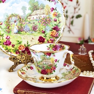 K&ouml;nnte beinhalten: Eine wei&szlig;e Teetasse und Untertasse mit einem Blumenmuster, das ein H&auml;uschen und Rosen zeigt. Die Teetasse und Untertasse haben einen goldenen Rand. Die Teetasse hat einen Henkel. Die Teetasse und Untertasse stehen auf einem roten Buch mit dem Titel "Adventures of Odysseus".